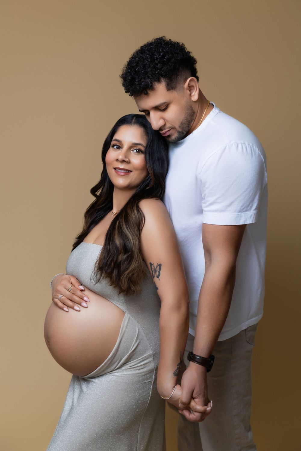 An intimate maternity portrait of an expectant couple embracing against a brown backdrop, wearing white and gray attire.