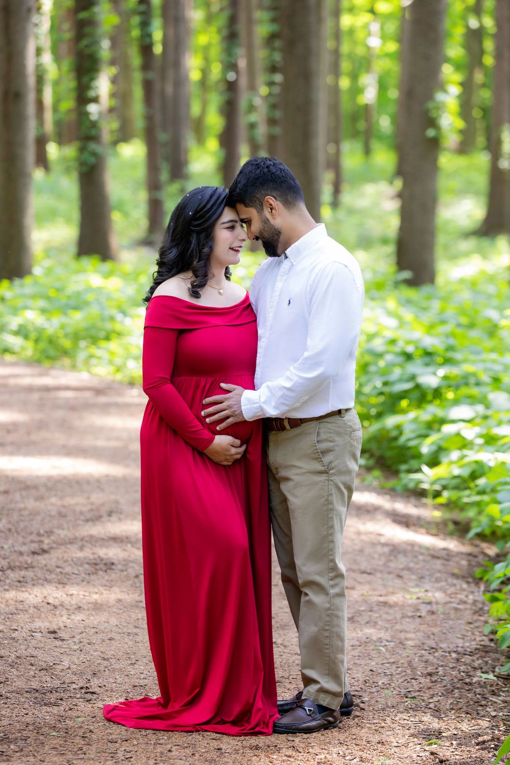 A romantic pregnancy photoshoot shows a couple embracing on a wooded path surrounded by lush green trees.