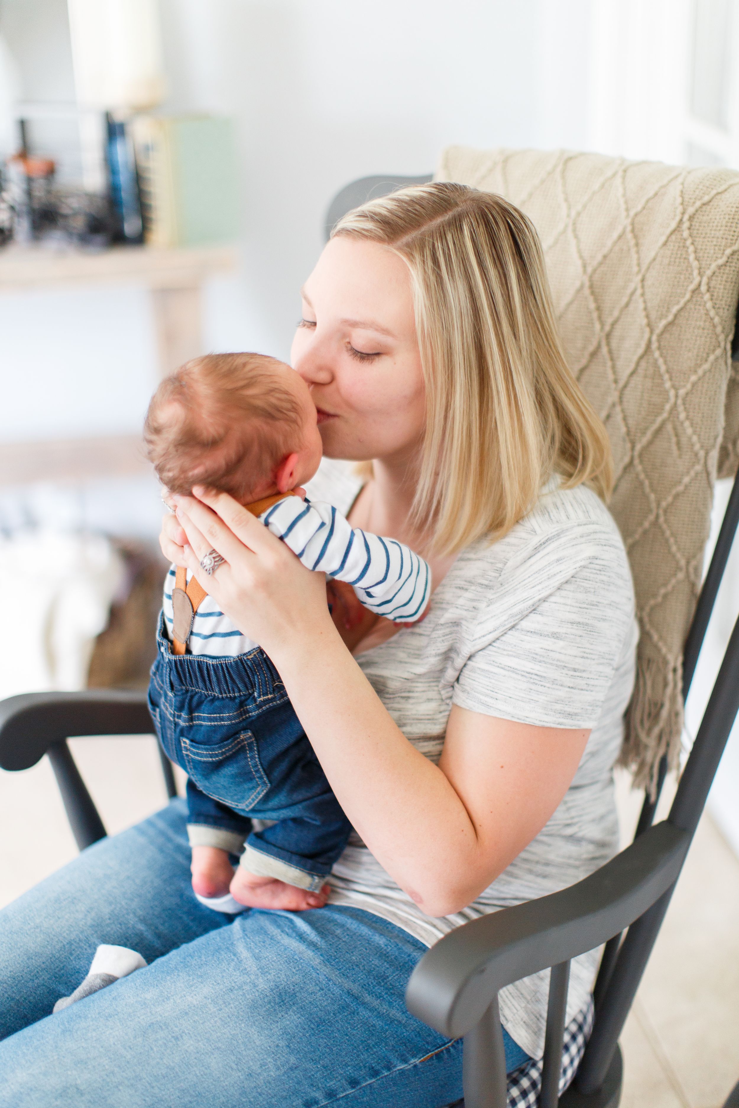 Intimate moment between parent and baby in casual wear sitting in grey rocking chair during a lifestyle newborn session in Springfield, MO.