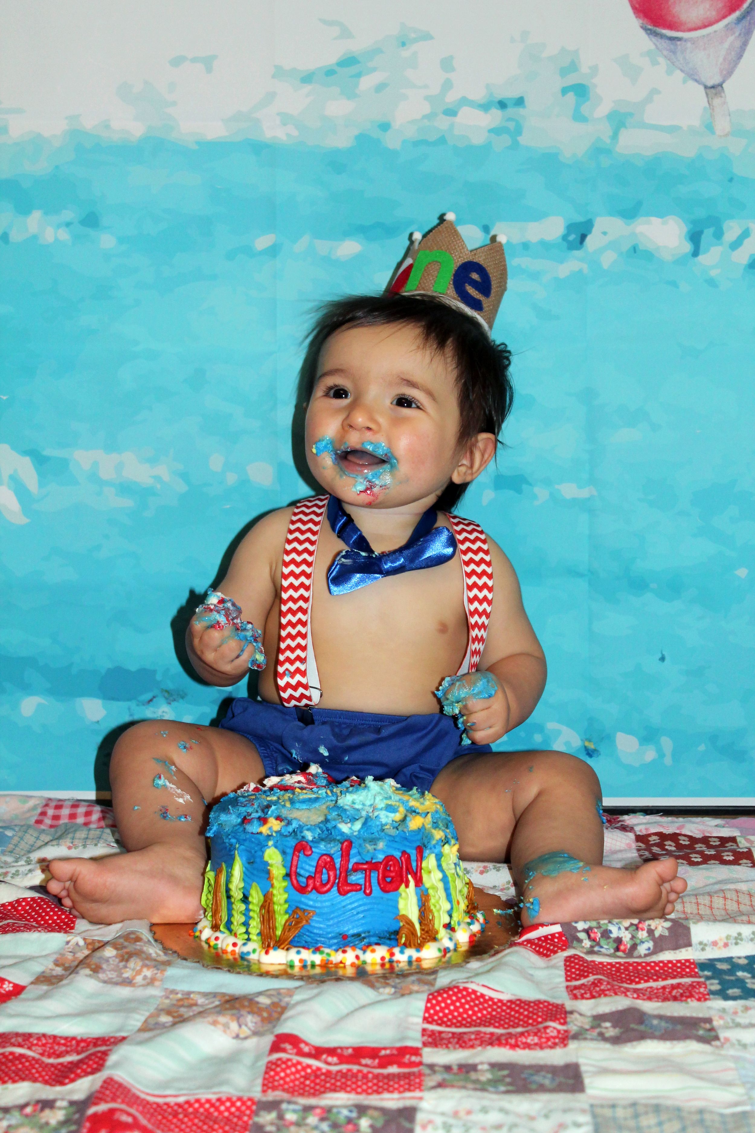 Baby wearing crown and suspenders celebrates first birthday with blue frosting covered cake.