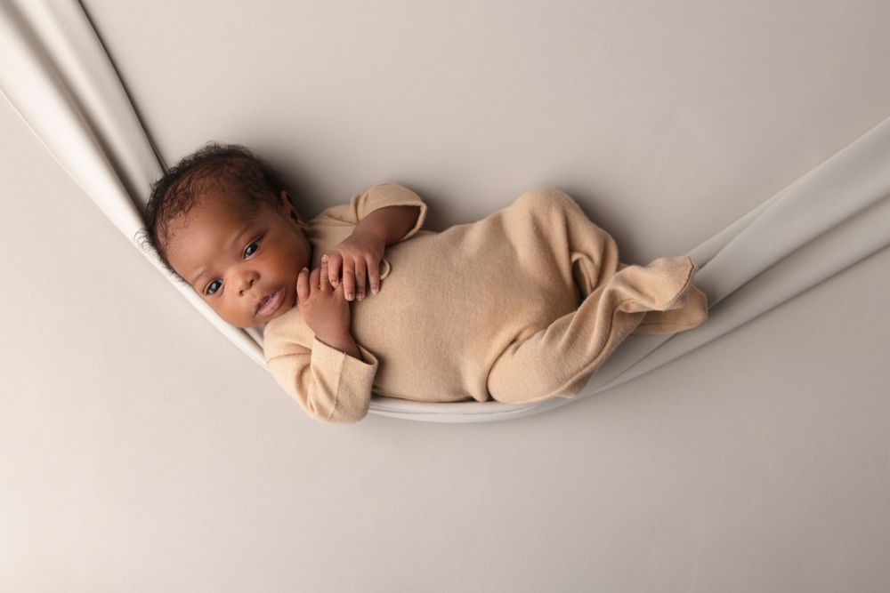 Sleeping baby wearing beige onesie resting in soft fabric hammock against white background.