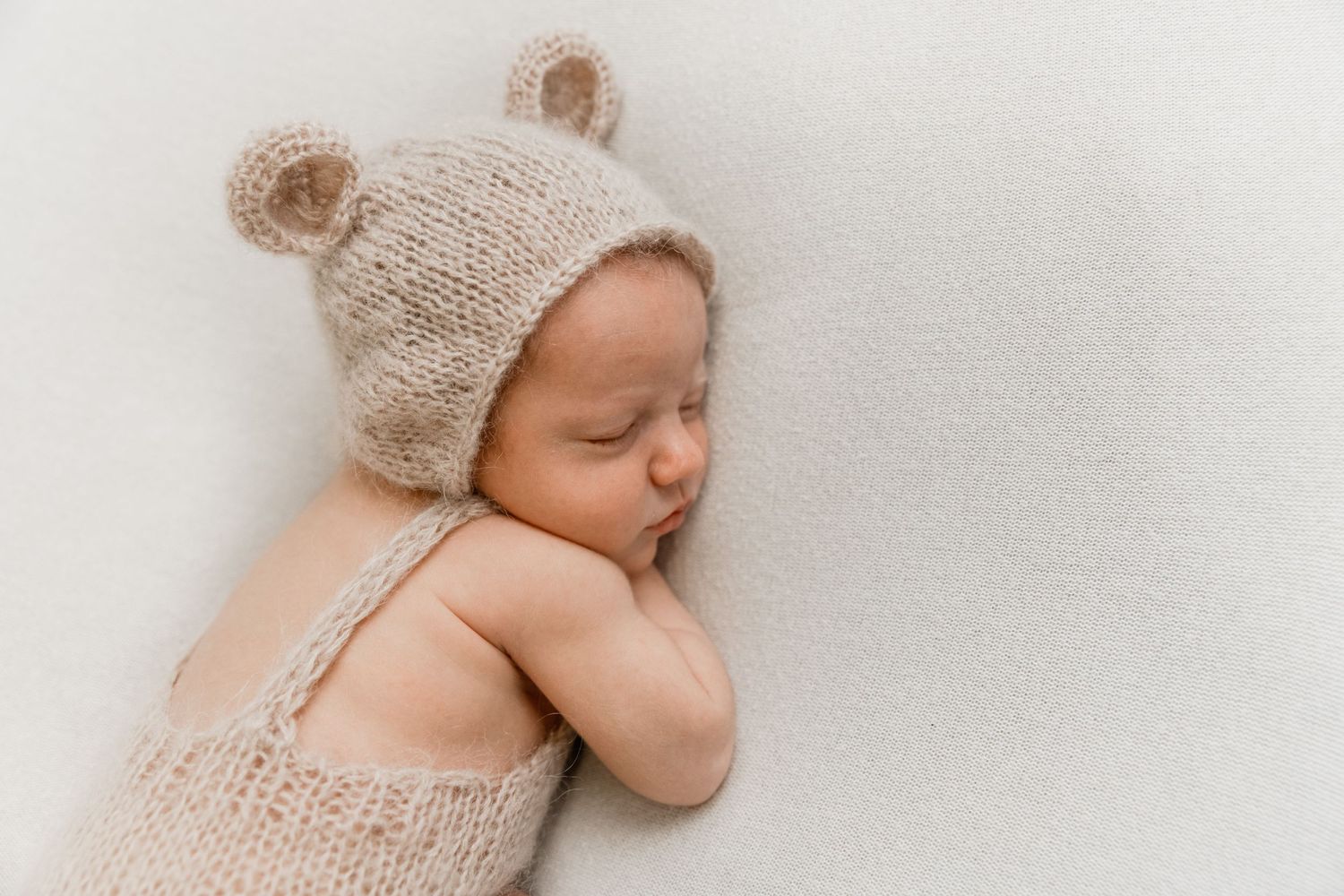 Sleeping newborn wearing a knit bear hat and romper in a minimalist setting.