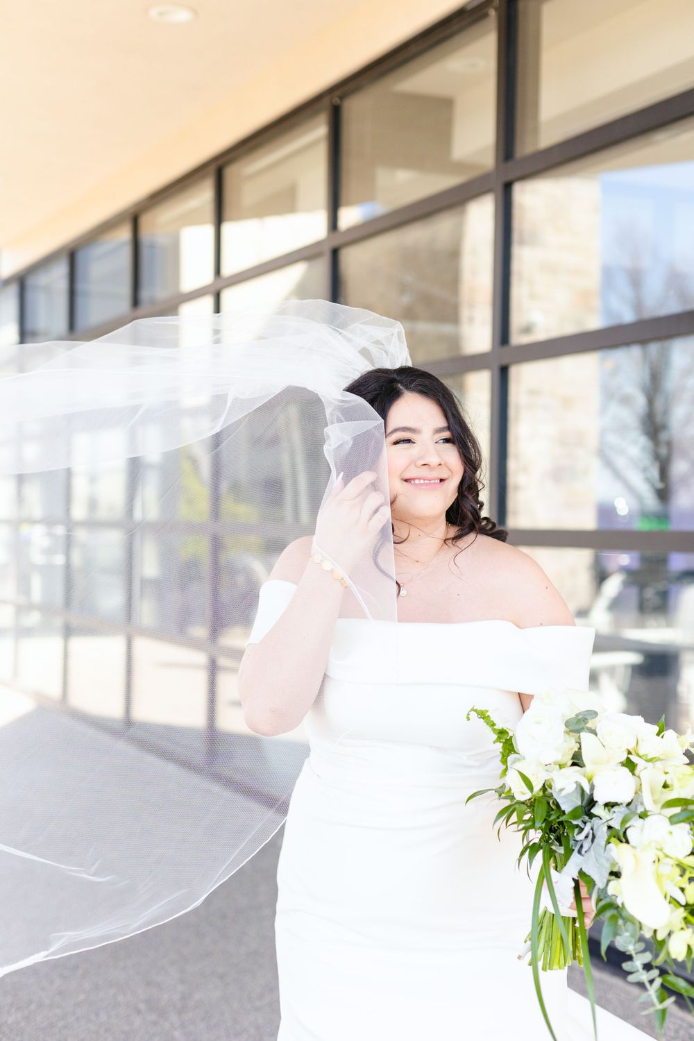 A bride in white dress with flowing veil smiles while holding a beautiful white floral bouquet.