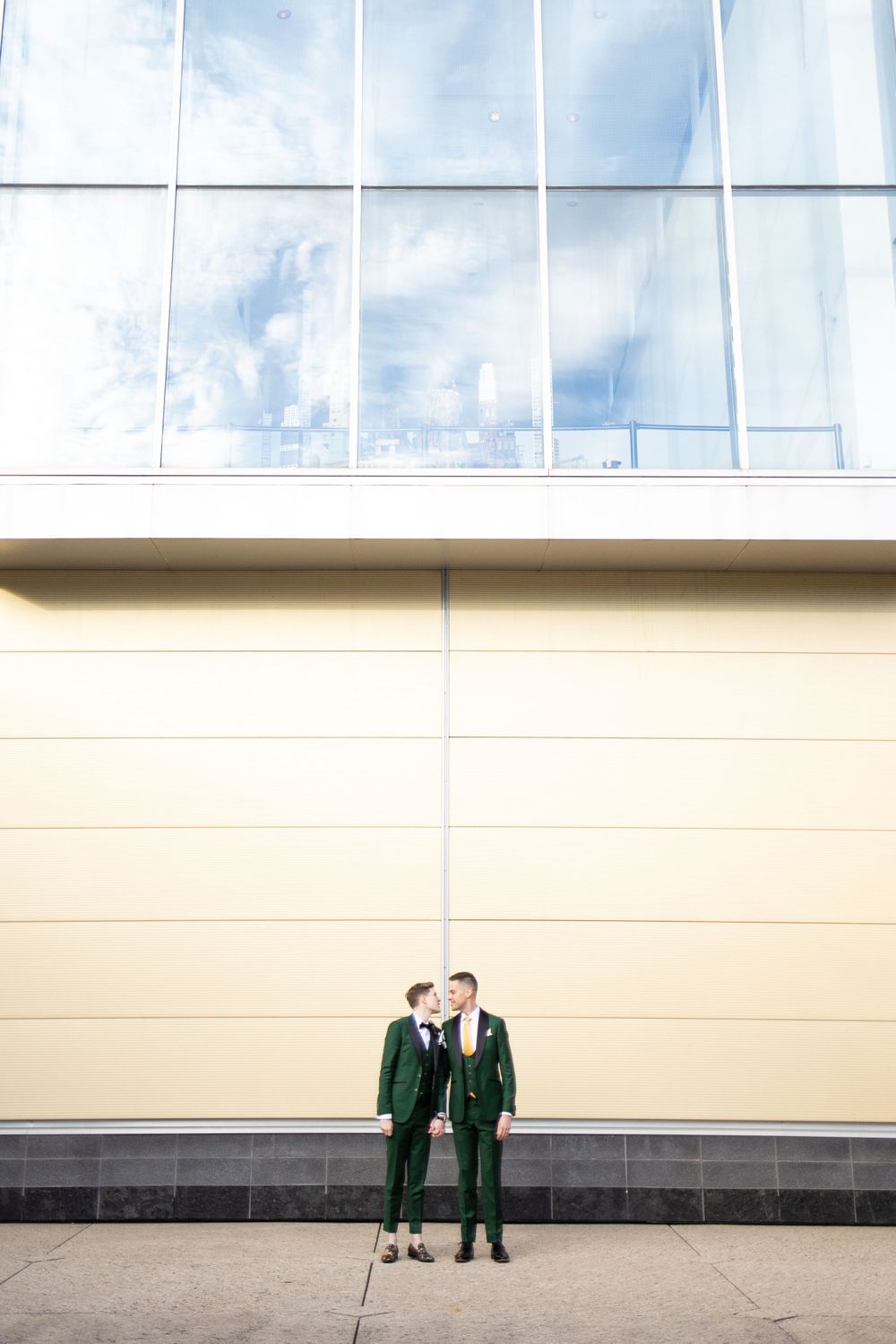 Wedding couple in green suits standing against modern building with glass windows.