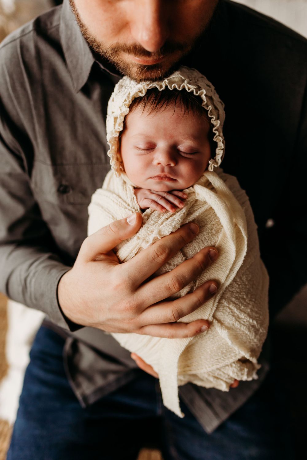 Newborn wearing vintage lace bonnet snuggled against caregiver in intimate portrait.