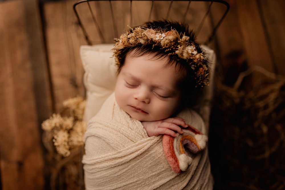 Sleeping newborn wearing dried flower crown wrapped in cream colored swaddle on wooden backdrop.