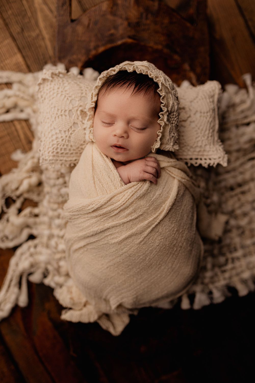 Peaceful sleeping newborn in cream wrap and bonnet on textured blanket with wooden background.