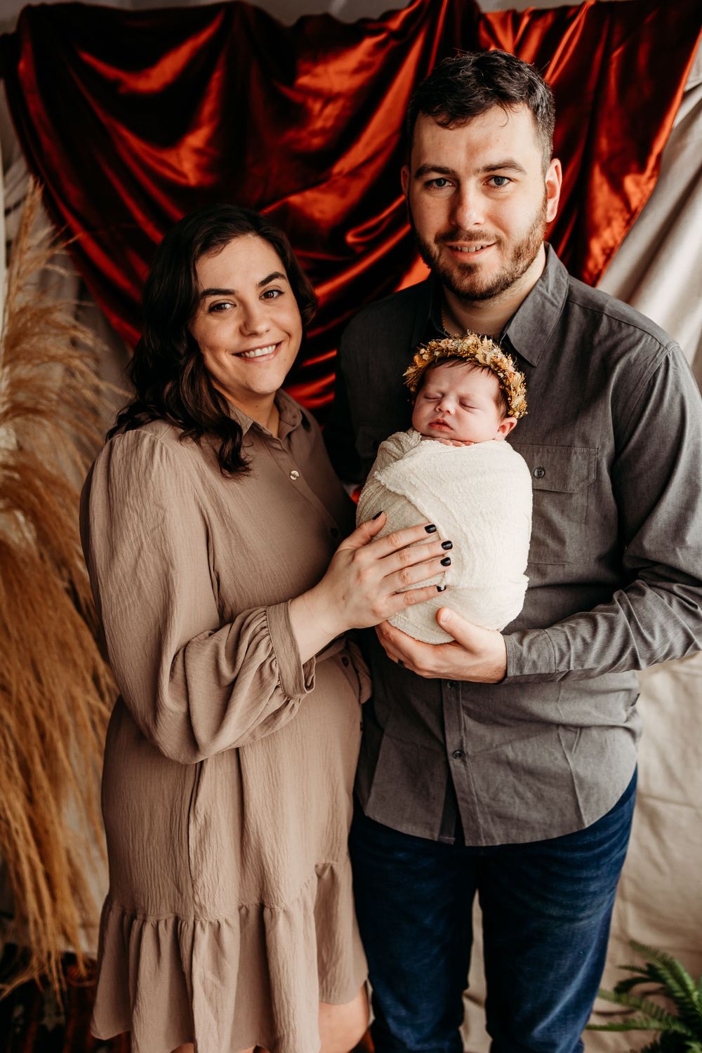 A family portrait with parents holding their newborn baby against a red fabric backdrop.