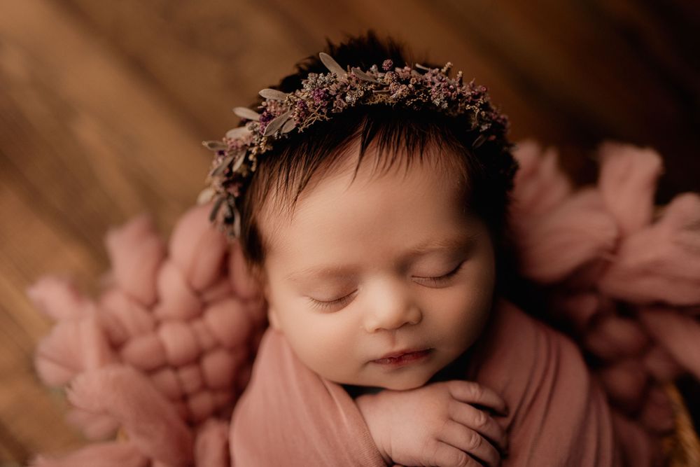 Sleeping newborn with floral crown wrapped in soft pink swaddle against dark wood background.