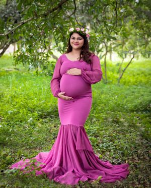 Outdoor maternity photo in pink mermaid dress with floral crown amidst green garden setting.