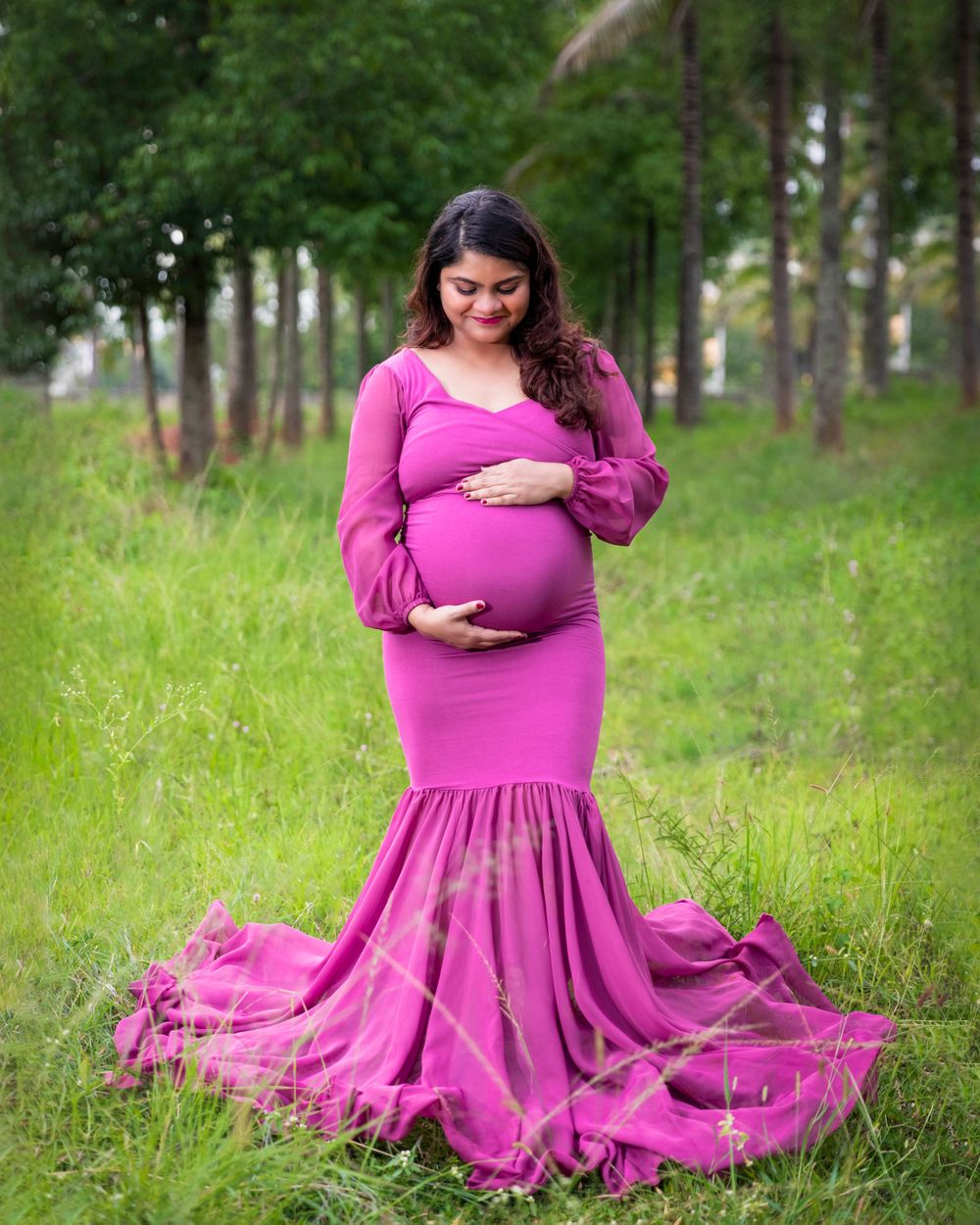 Expectant mother in flowing pink gown poses in forest clearing.