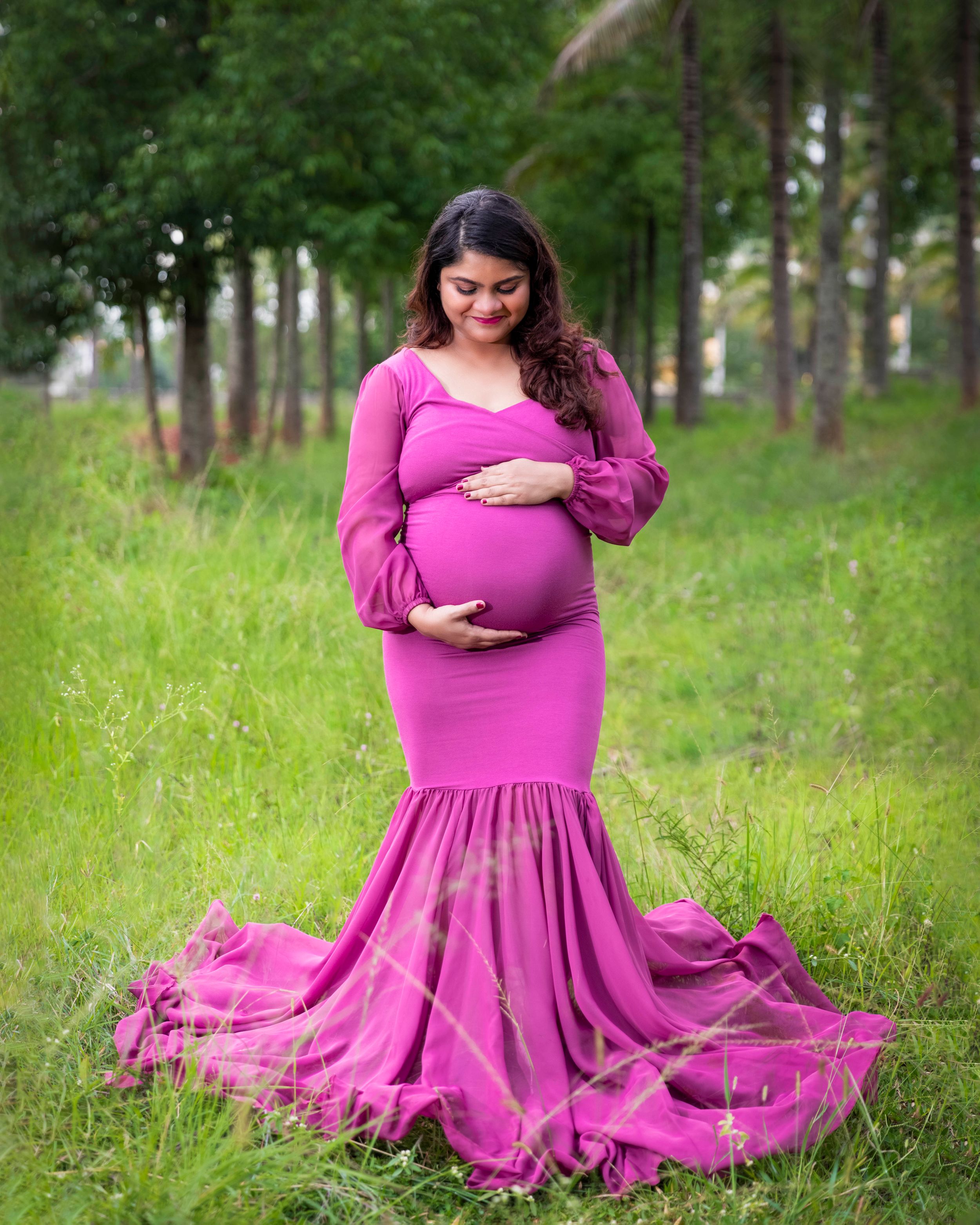 Expectant mother in flowing pink gown poses in forest clearing.