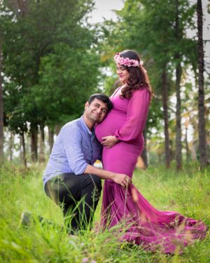Outdoor maternity photoshoot featuring couple in pink dress and blue shirt among trees.