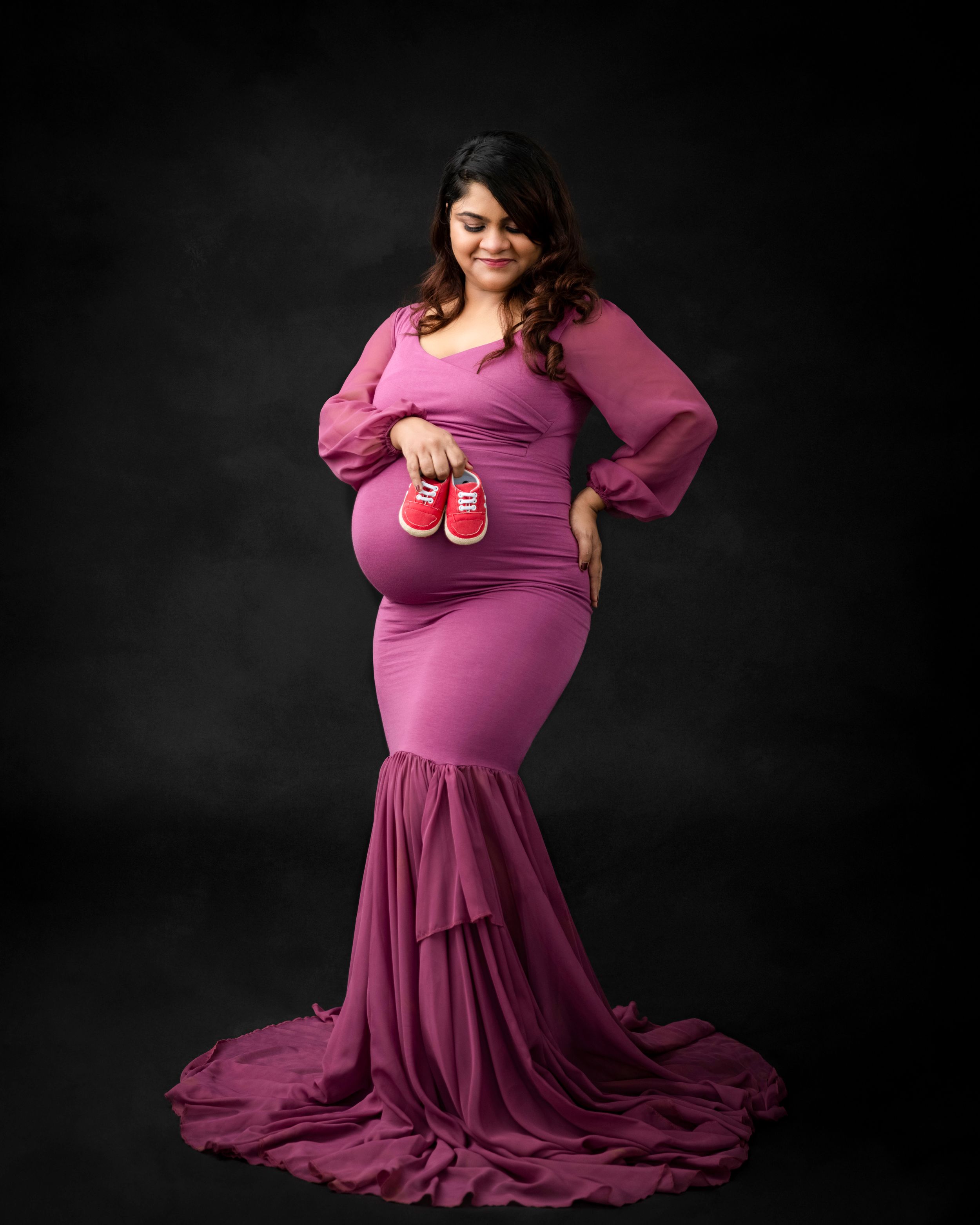 Studio maternity portrait with tiny red baby shoes against dark background.