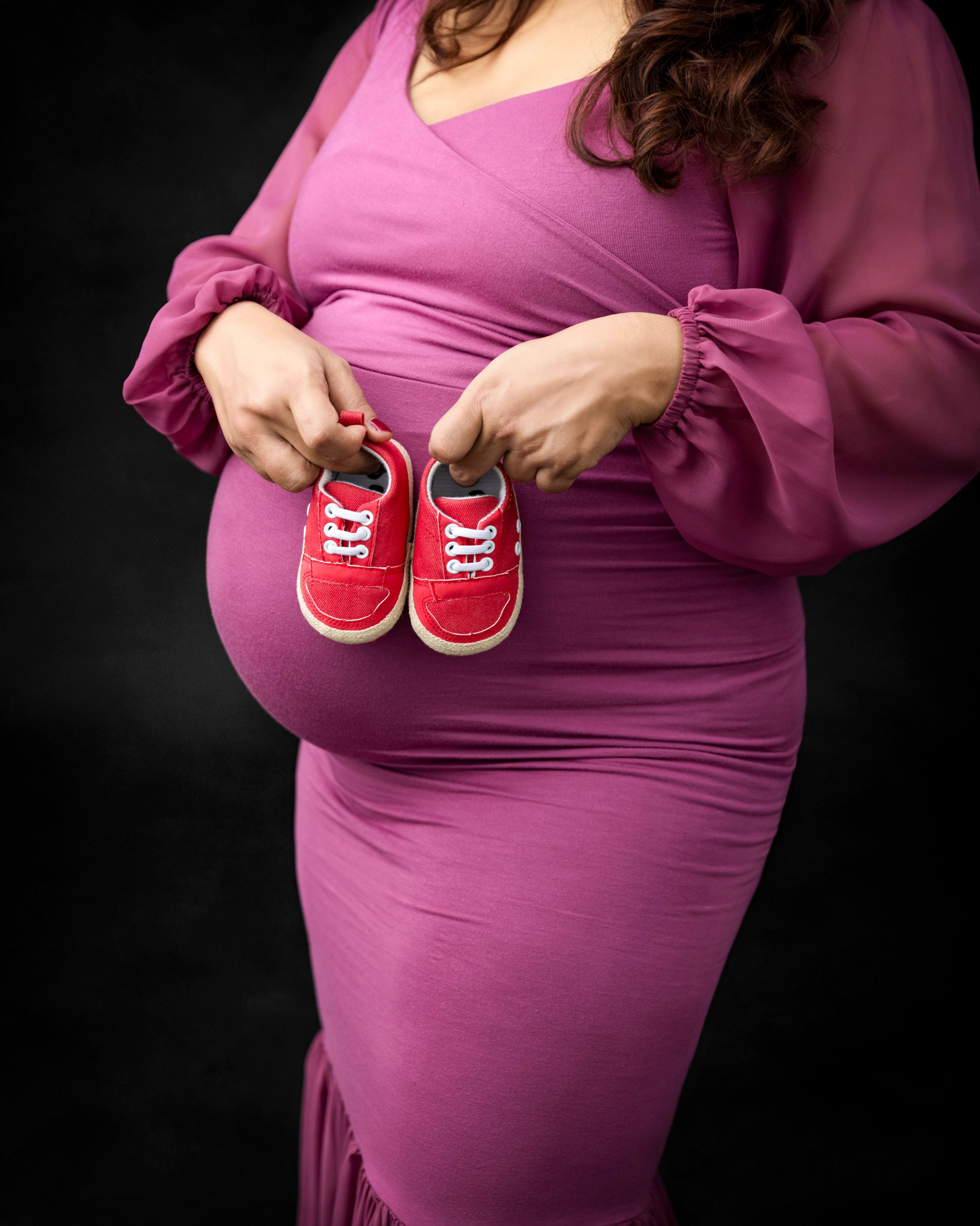 Close-up maternity photo showing red baby shoes held against pink dress.