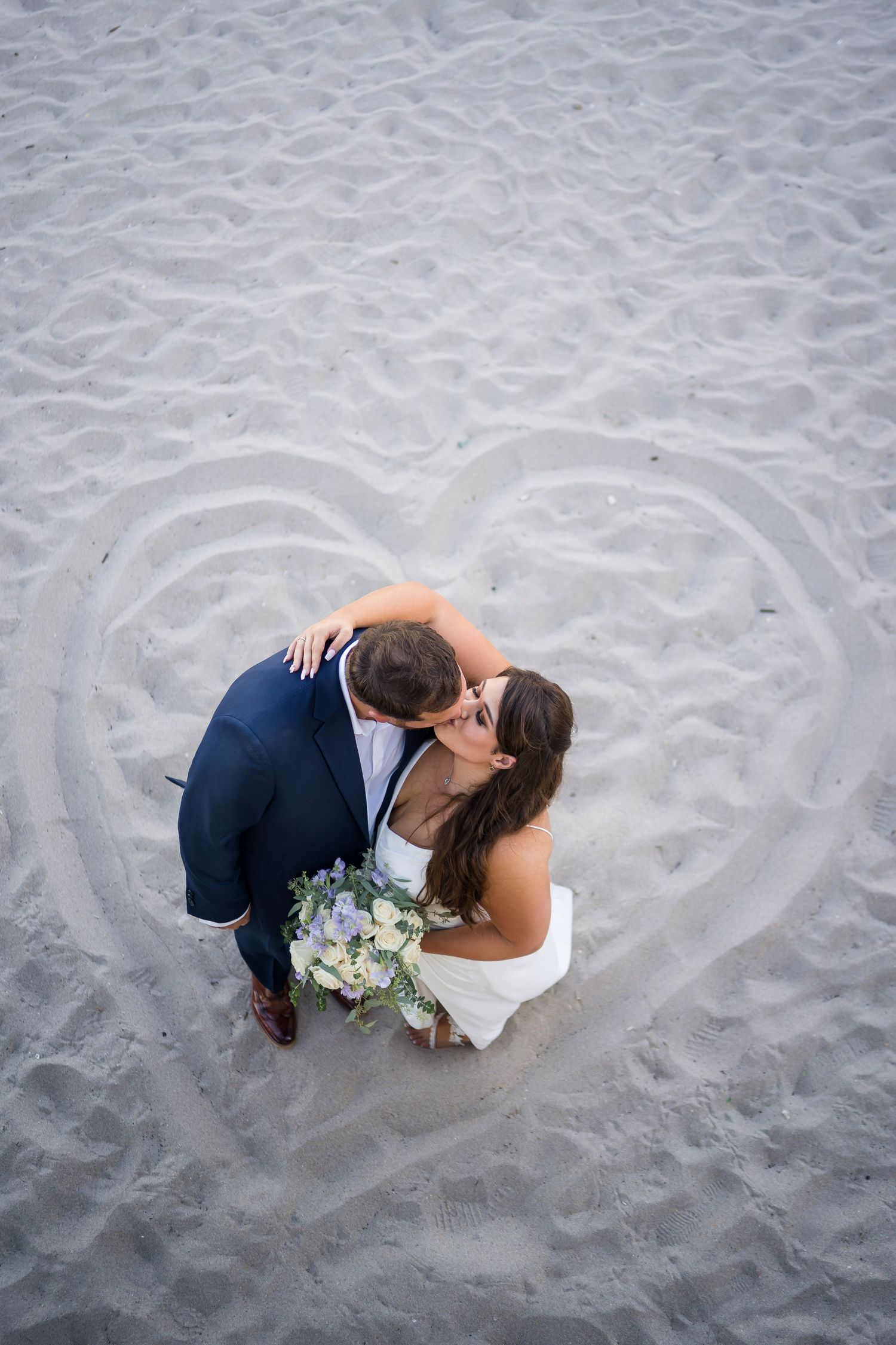 Aerial view of newlyweds embracing on beach with heart drawn in sand and blue flower bouquet.