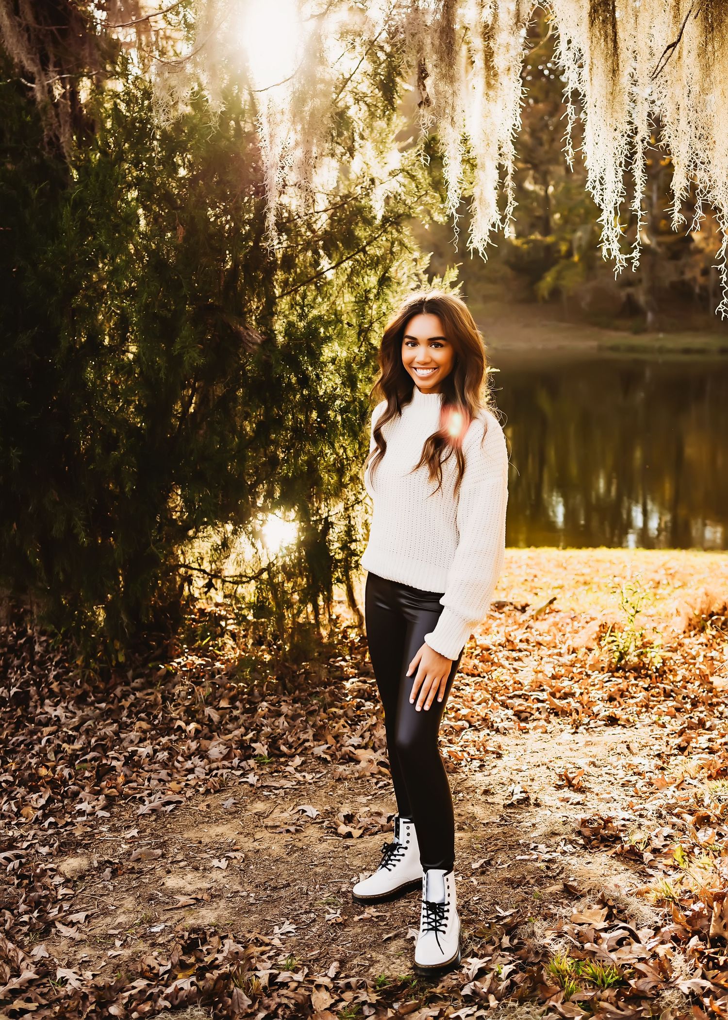Person in white sweater standing by mossy lake with sunlit Spanish moss.