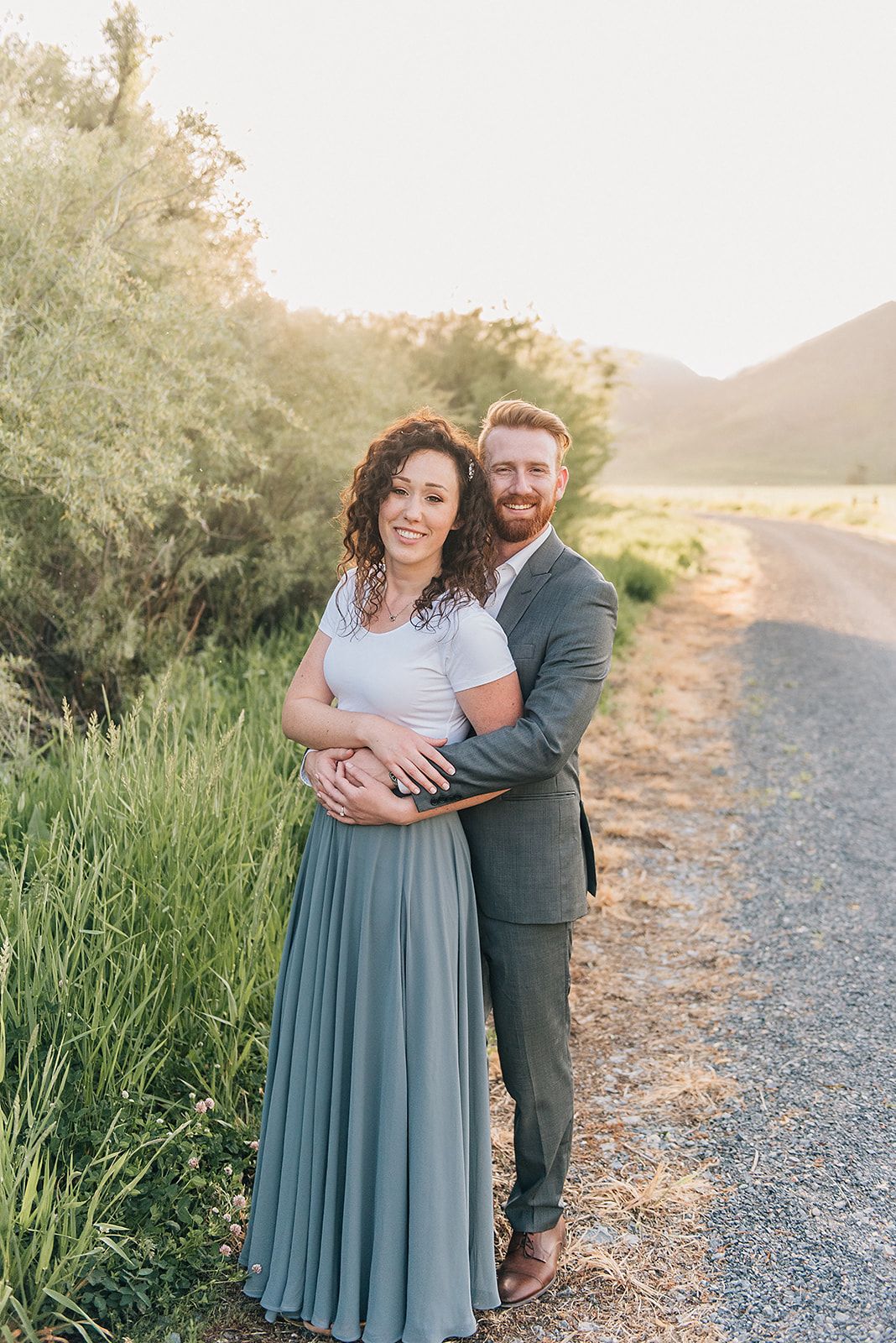 A couple embracing on a dirt path with mountains and sunset in the background.
