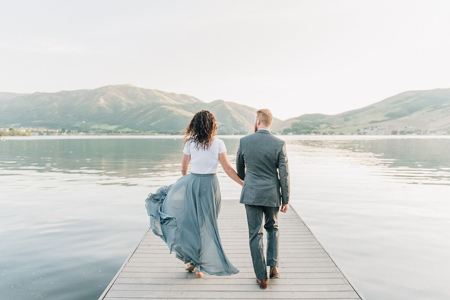 A couple walking hand in hand on a wooden pier overlooking a serene lake.