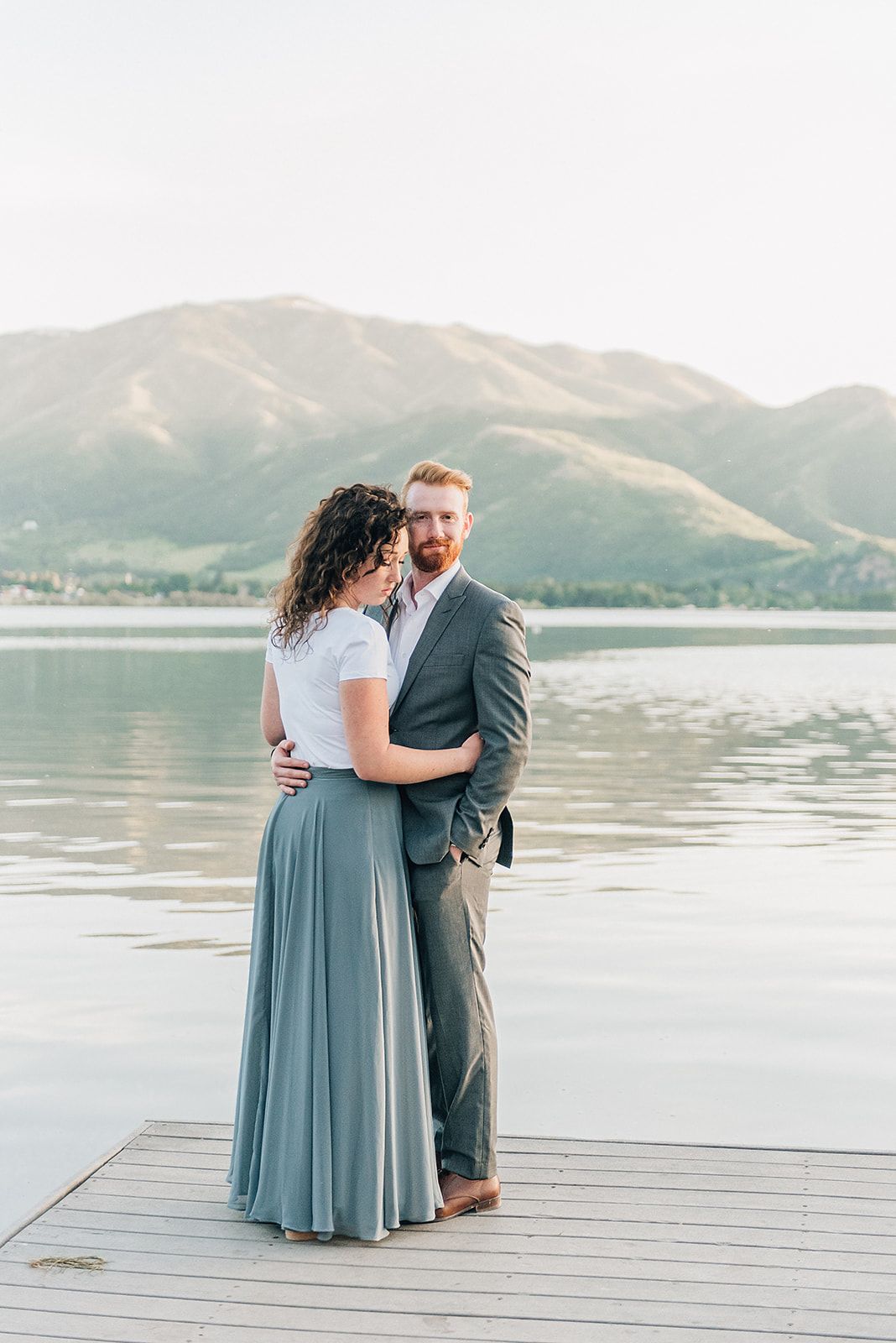 A couple in formal attire embracing on a dock with mountains and lake in the background.