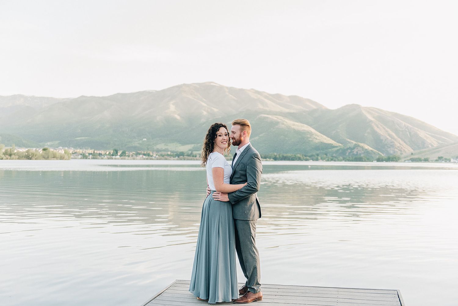 A couple embracing on a lakeside dock with mountains in the background.