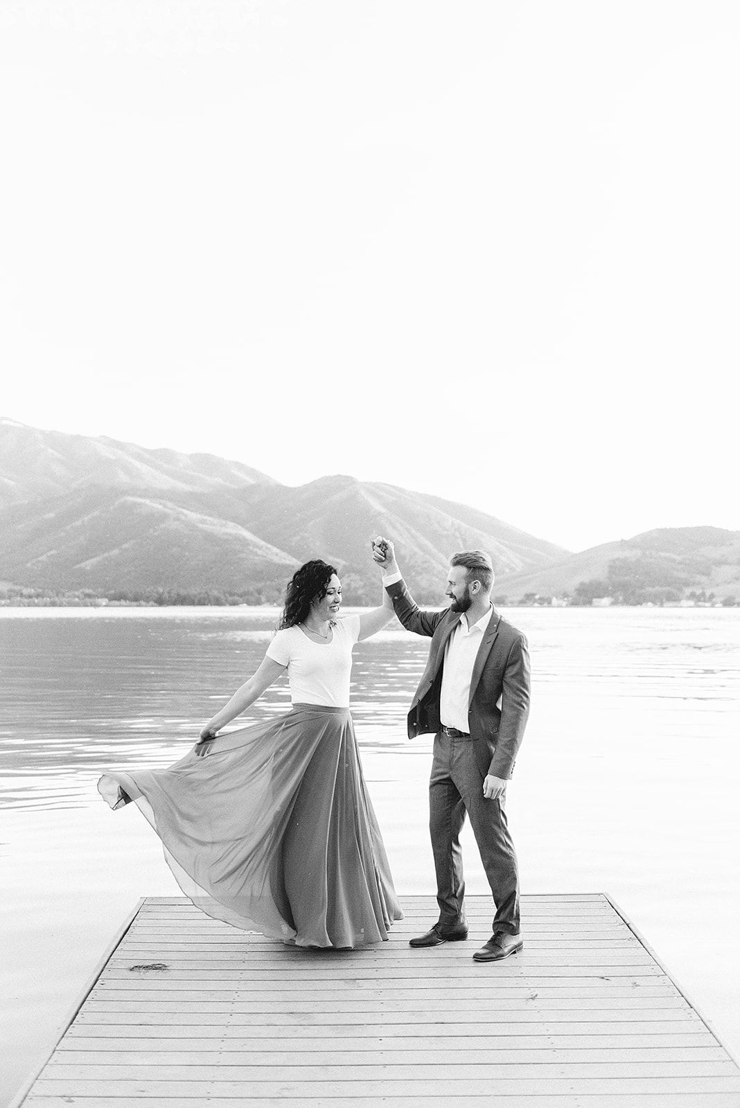 A couple dancing on a wooden dock with mountains and lake in the background.