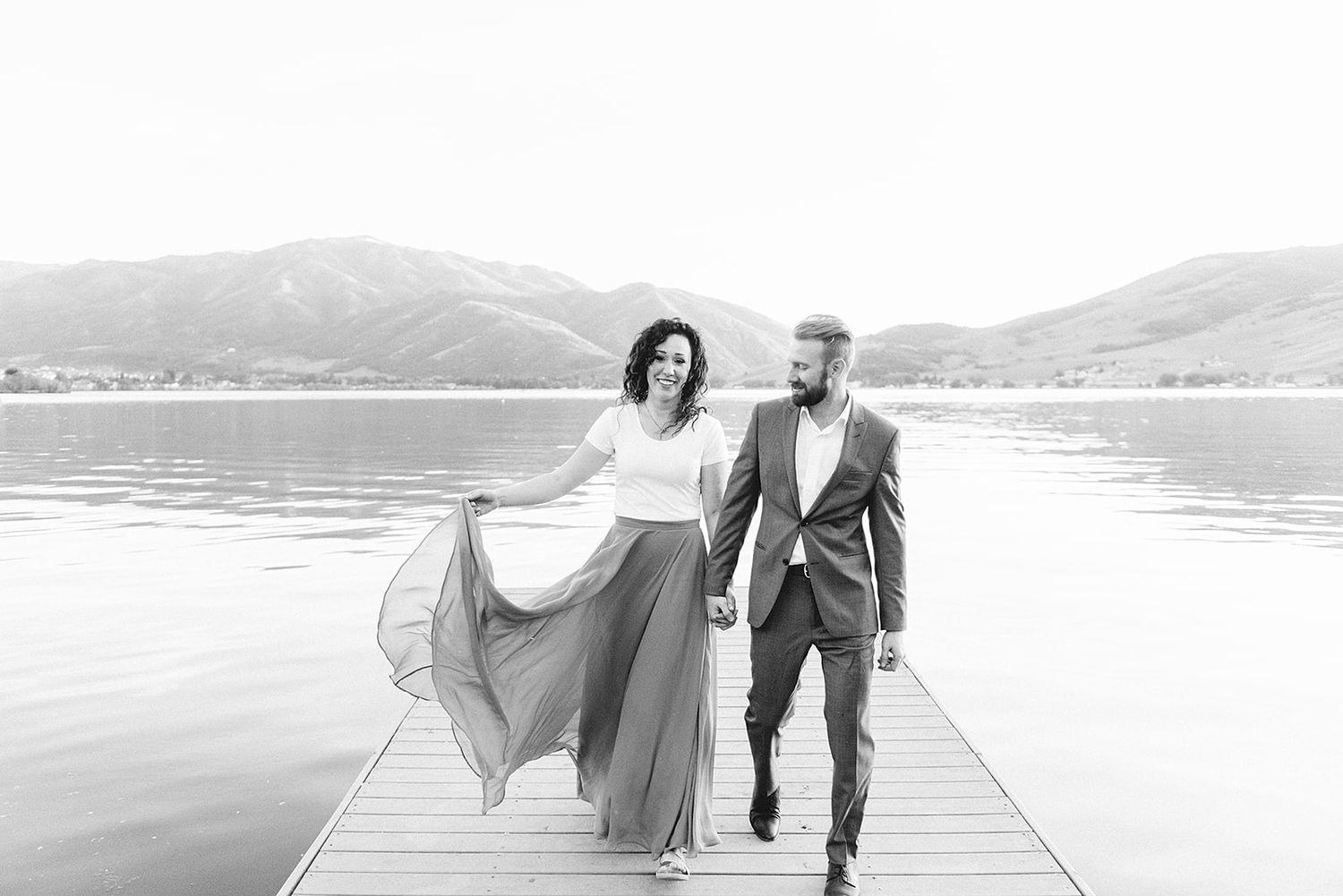 A couple walking on a wooden dock by a lake with mountains in the background.