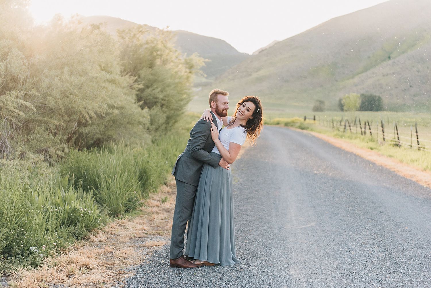 A couple sharing an intimate moment on a country road with mountains and greenery.