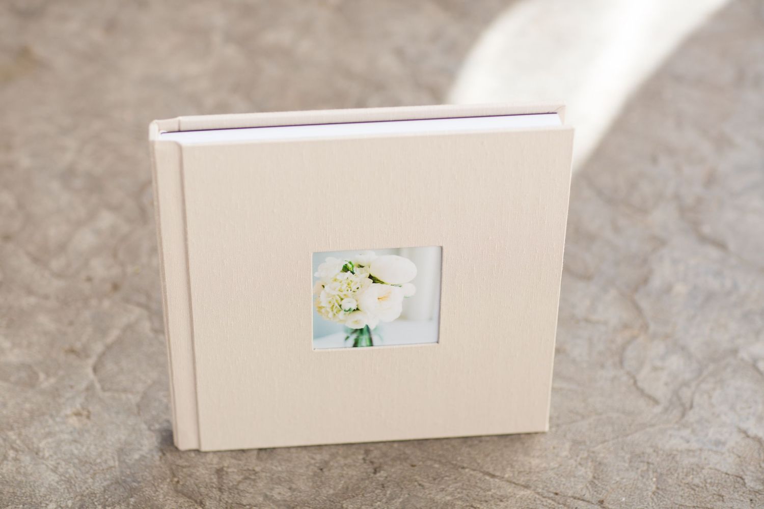 A cream-colored photo album with a small floral insert lies on a textured stone surface.