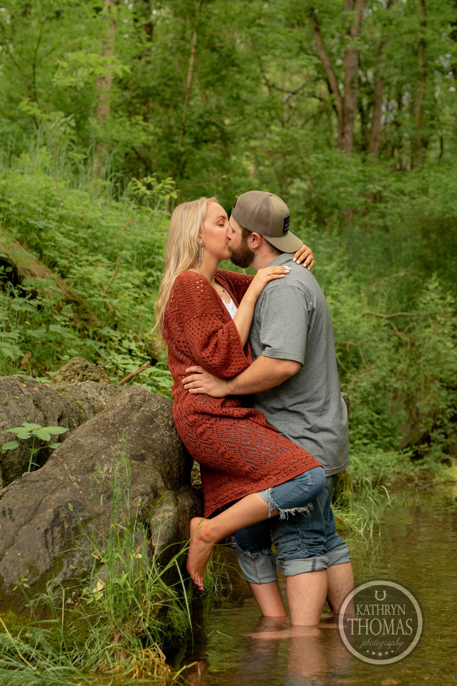 A romantic couple embraces while standing in a shallow creek surrounded by green forest.