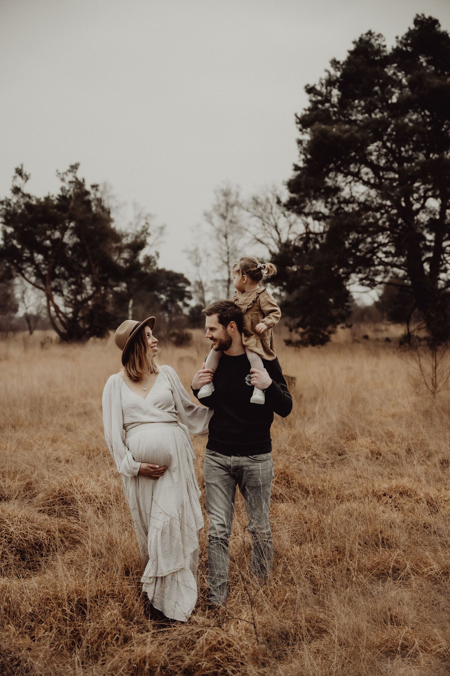 Family standing together in golden field during sunset with expectant mother in flowing dress.