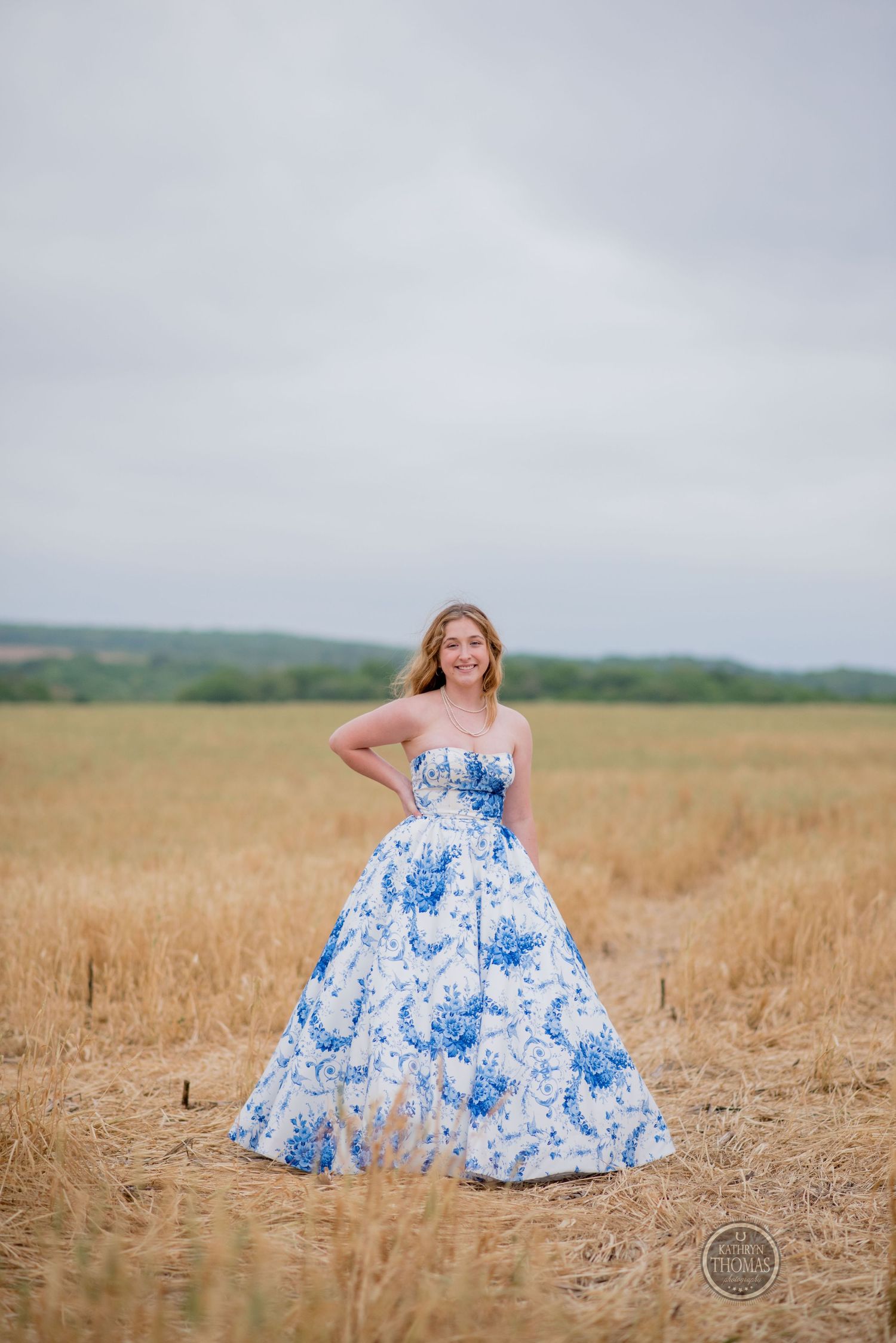 Model in a flowing blue and white floral ball gown poses gracefully in a golden wheat field under cloudy skies.