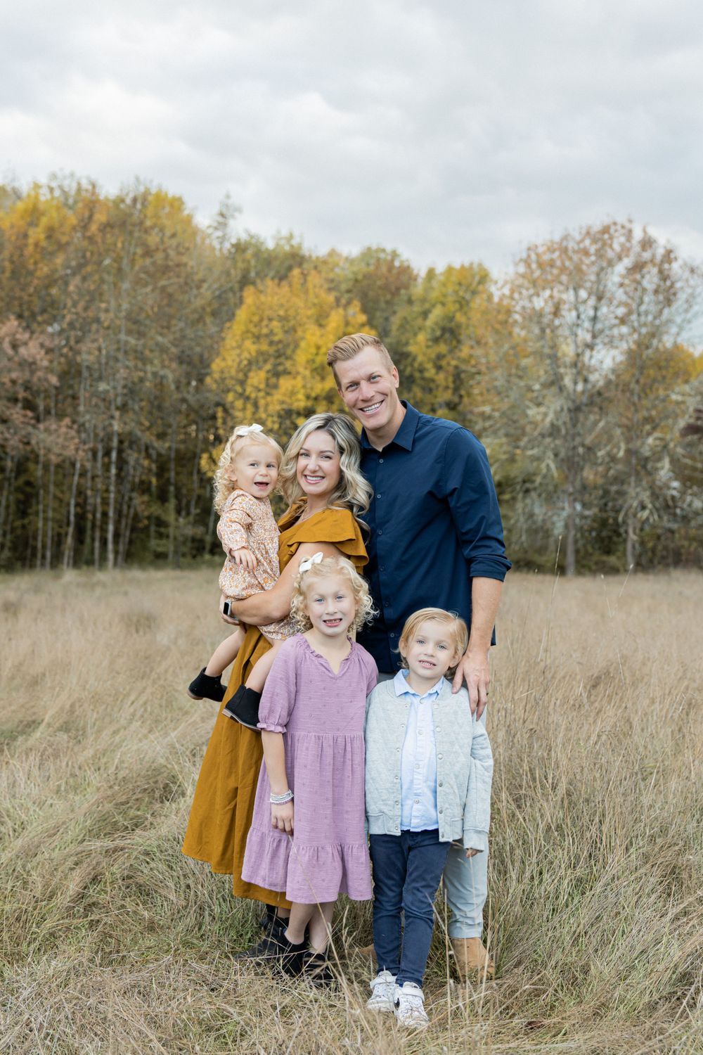 A family poses together in a grassy field with autumn trees featuring golden foliage in the background. Fleck of light photography.