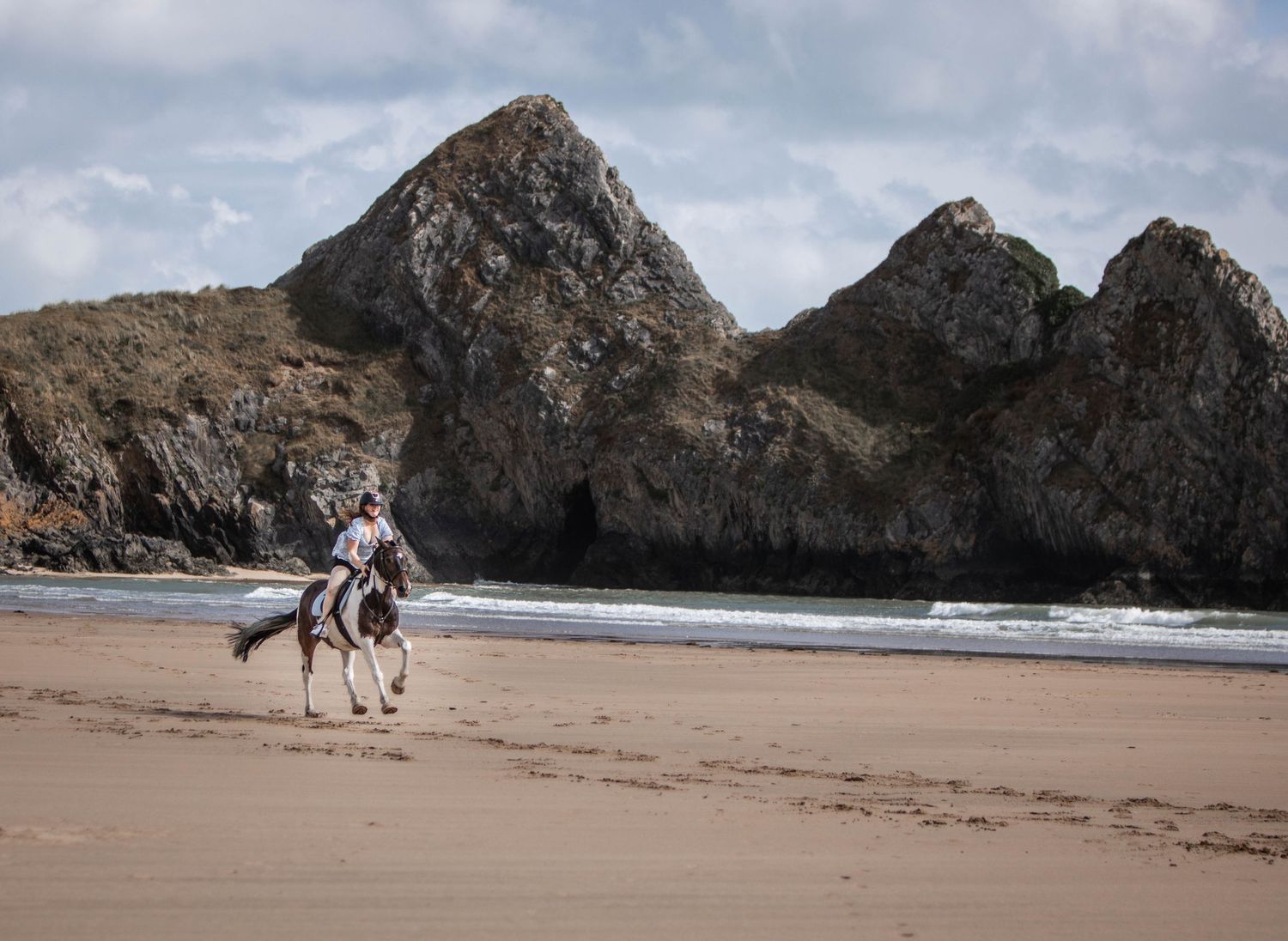 A horse and rider traverse a dramatic beach landscape with towering rocky cliffs.