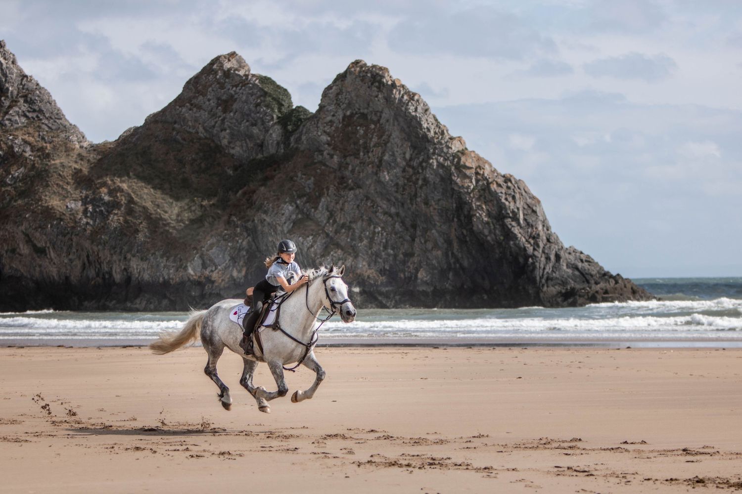 A white horse gallops past majestic coastal rock formations on a scenic beach.