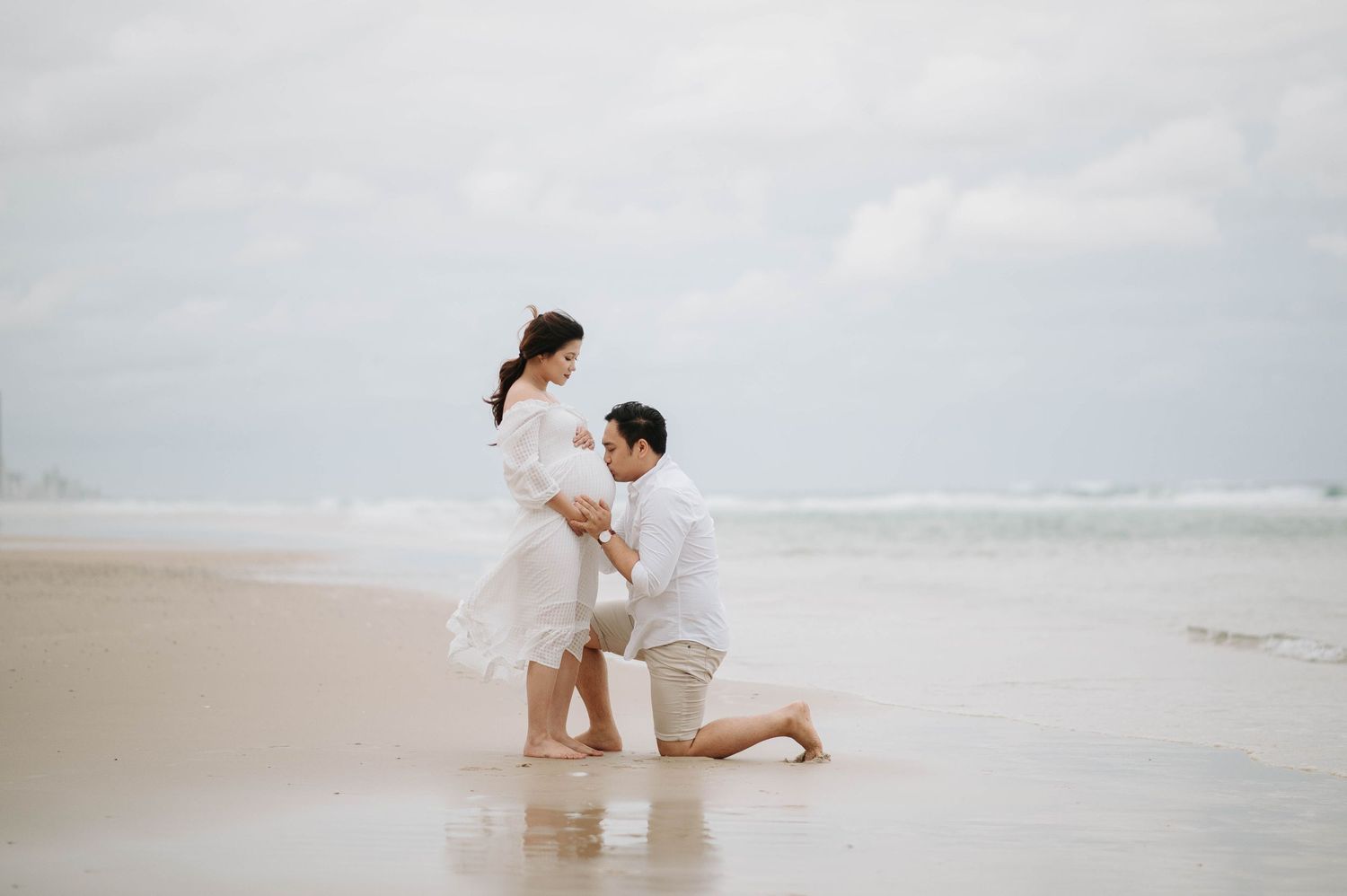 A romantic beach proposal scene with white dress and khaki shorts against an overcast ocean backdrop.