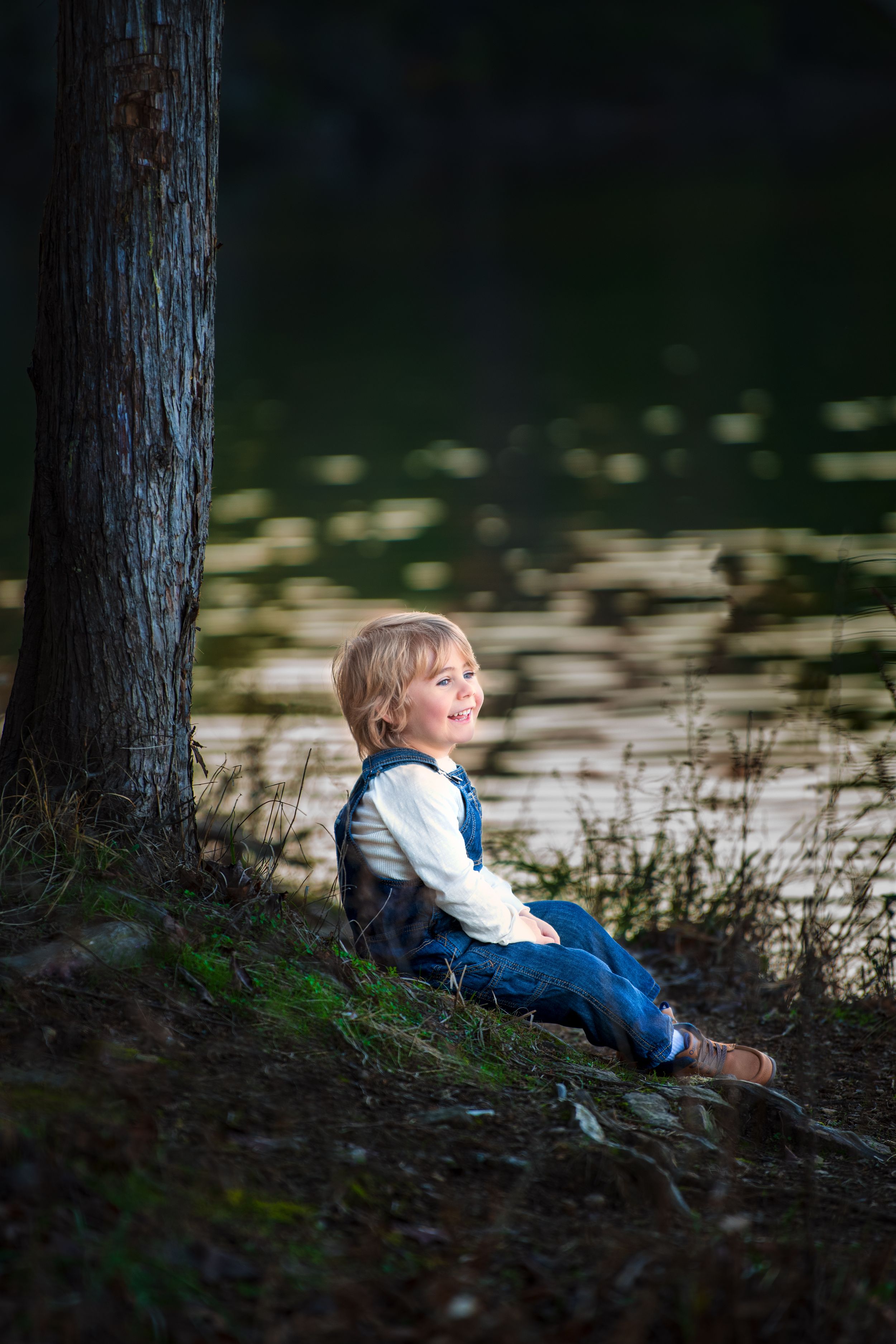 Small boy sitting on the edge of a lake wearing a white longsleeve shirt and denim overalls.