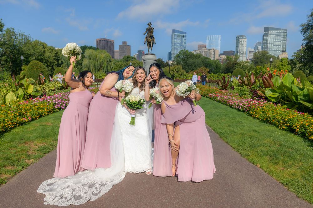 A bridal party in pink dresses poses in a garden path with city skyline visible in the background.