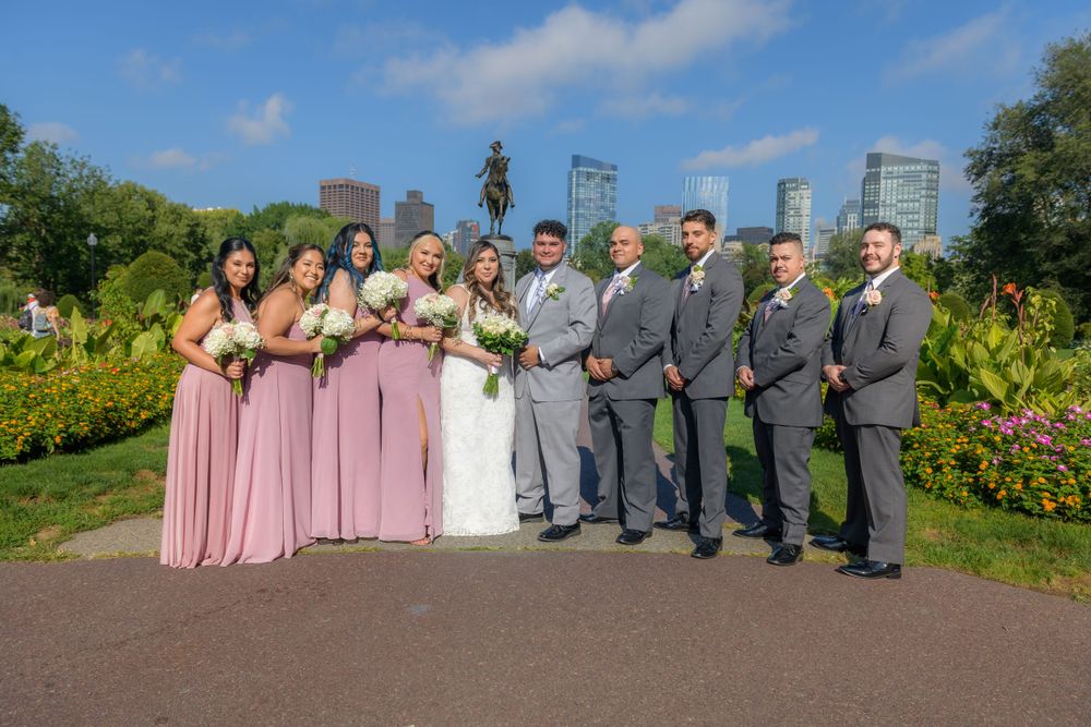 Wedding party in formal attire poses outdoors in a garden setting with pink bridesmaids dresses and gray suits.