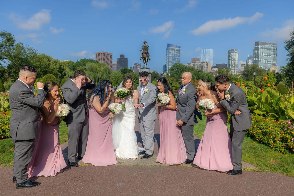 Wedding party in pink and gray attire poses together in a garden setting with city skyline in background.