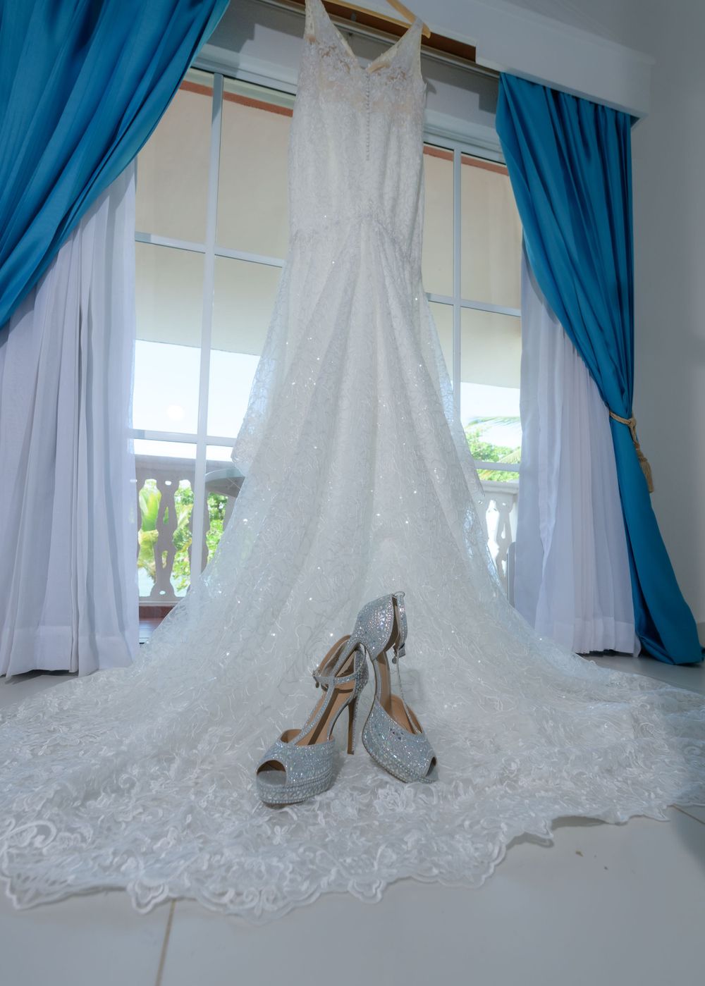 A white lace wedding dress hangs elegantly by a window with blue and white curtains, with silver heels displayed below.