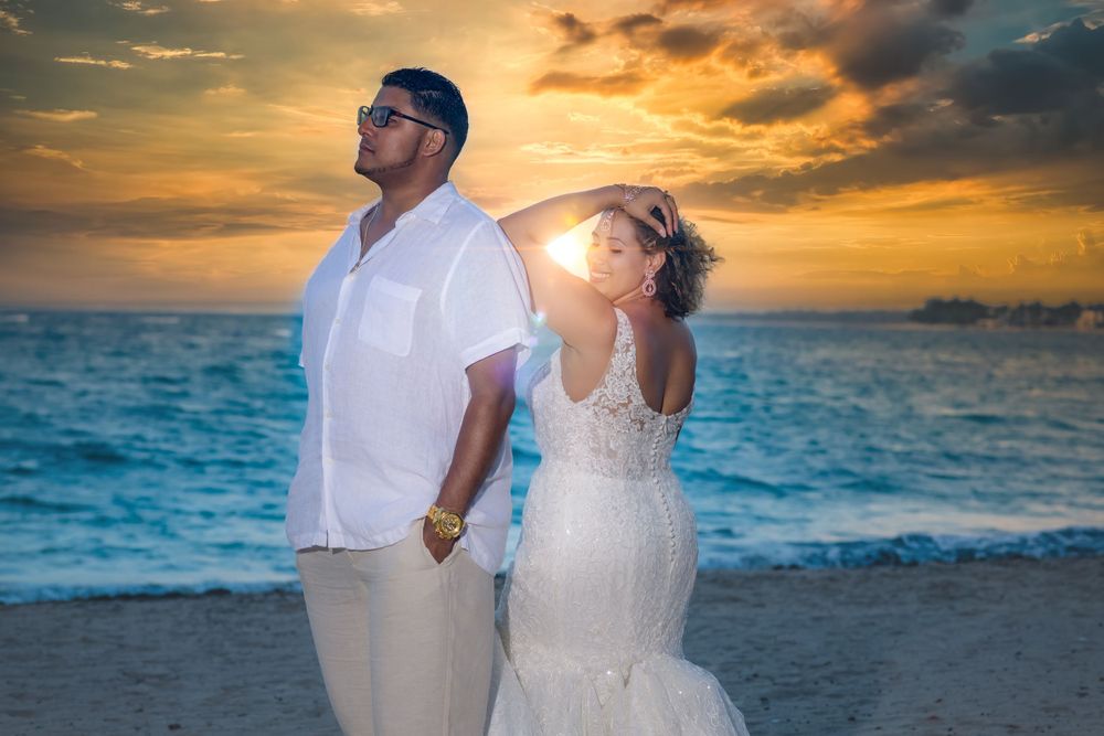 A couple in white attire poses back-to-back on a beach at sunset with dramatic orange clouds and turquoise waters.