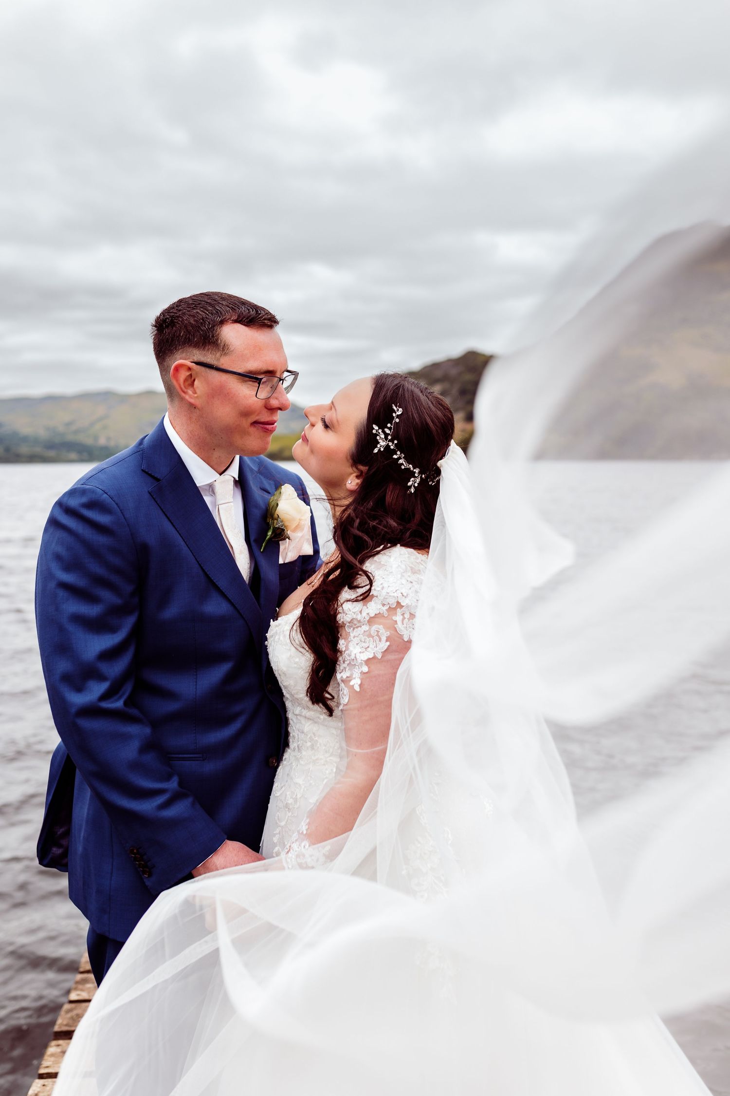 A wedding couple shares an intimate moment on a wooden pier with flowing veil in the wind.