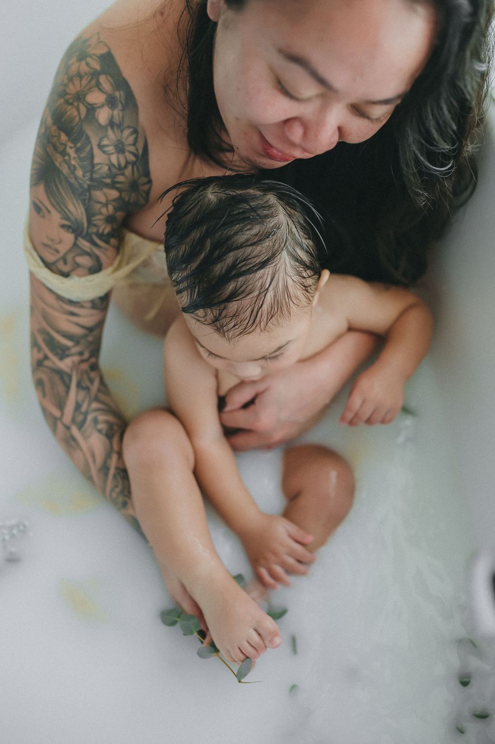 Tender bath time moment showing tattoo-adorned parent washing small baby in a milky white bath.