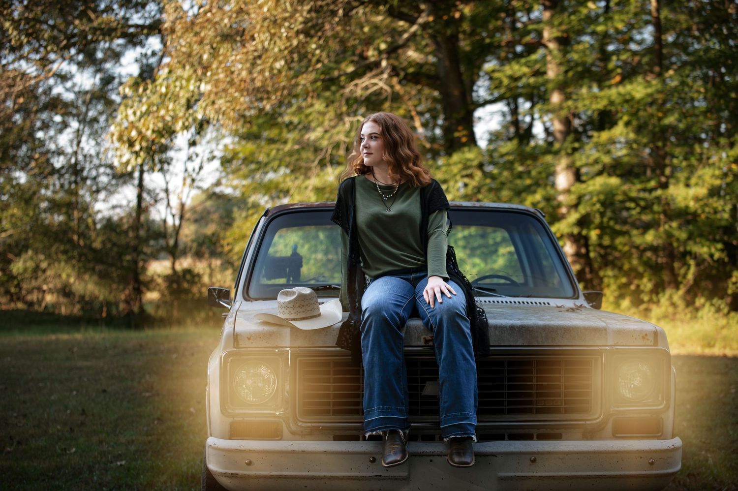 Person in casual attire sits on hood of vintage truck in autumn setting.