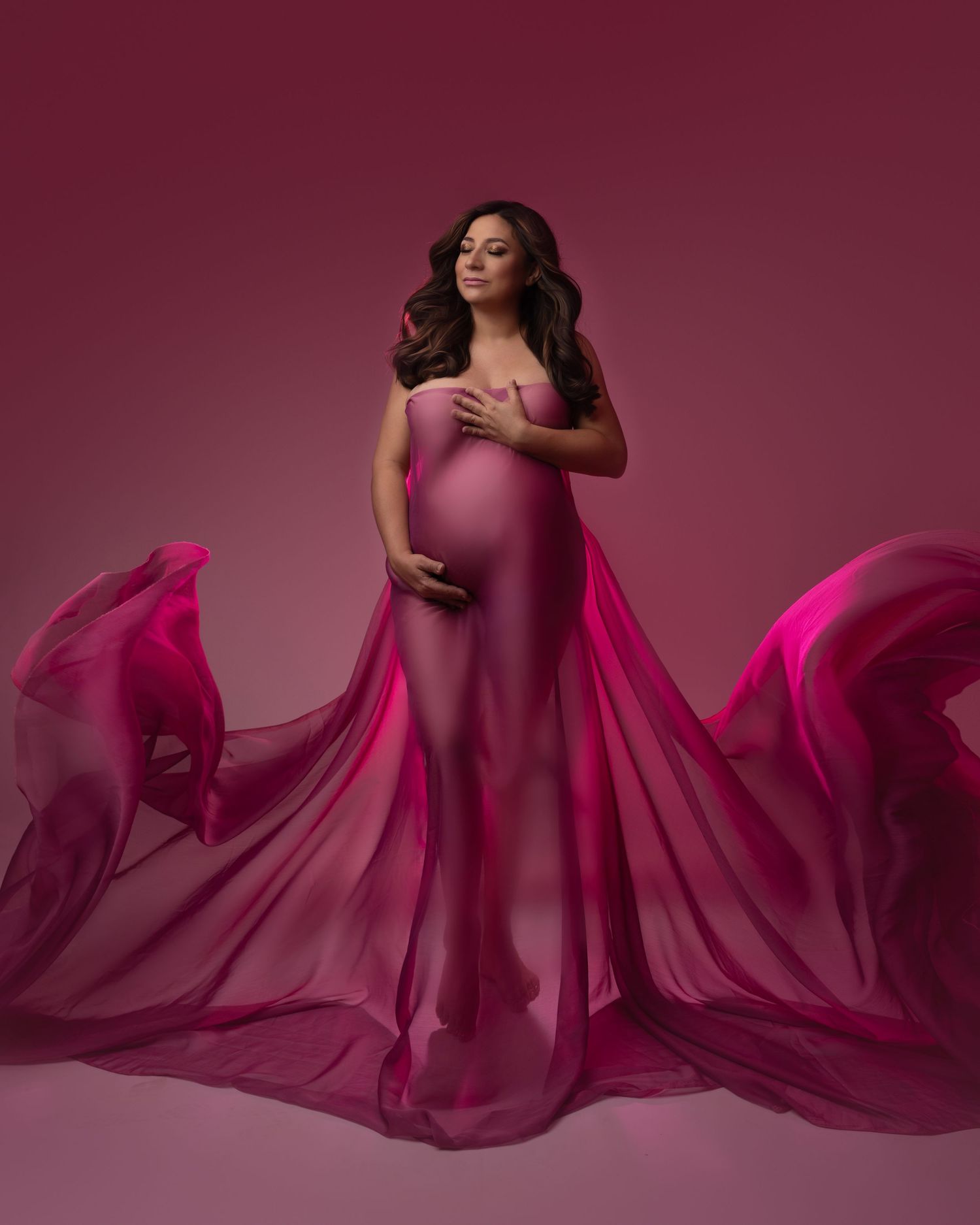 Elegant maternity photoshoot featuring flowing pink fabric against burgundy backdrop.