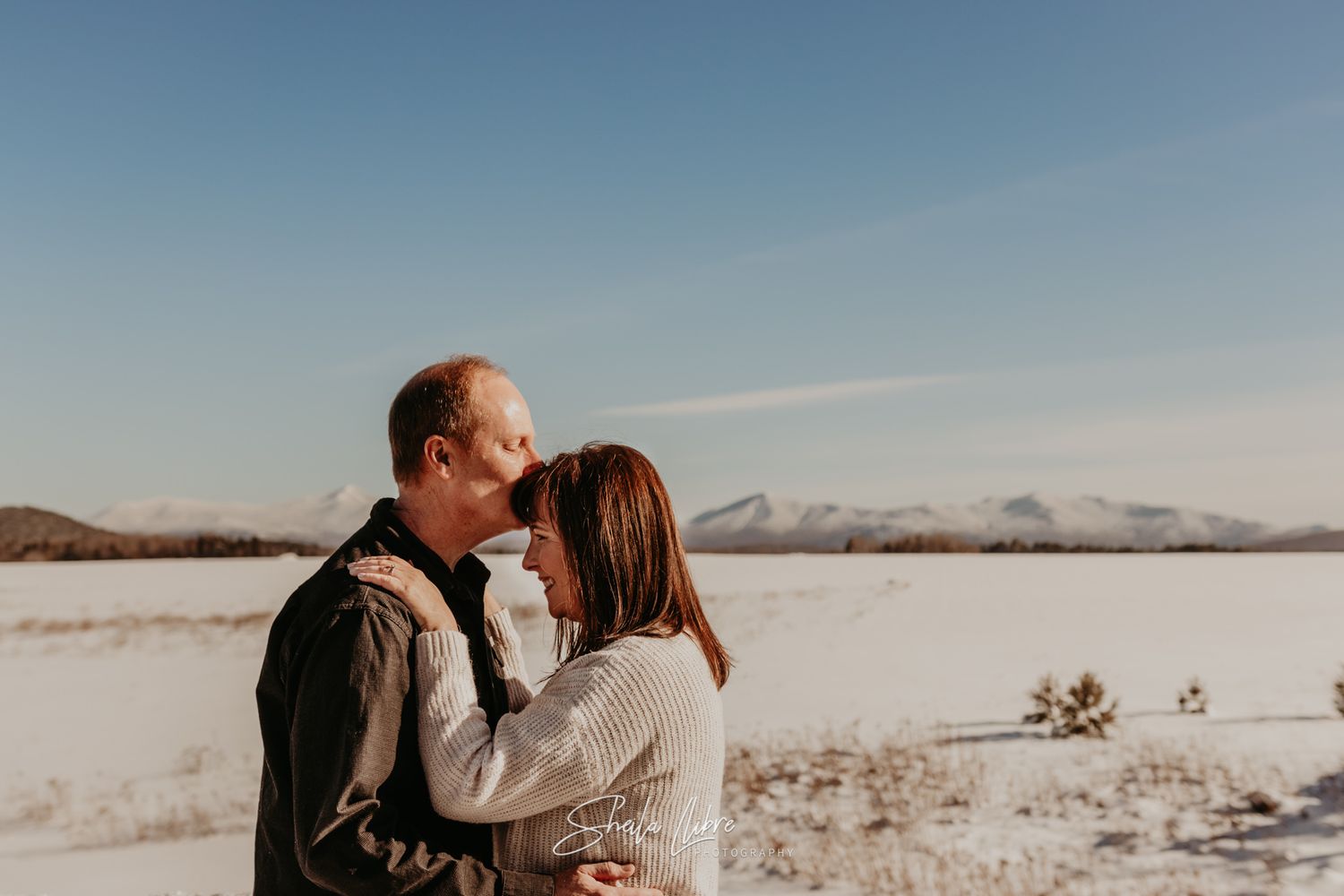 Couple embracing in winter landscape with snowy mountains in background.