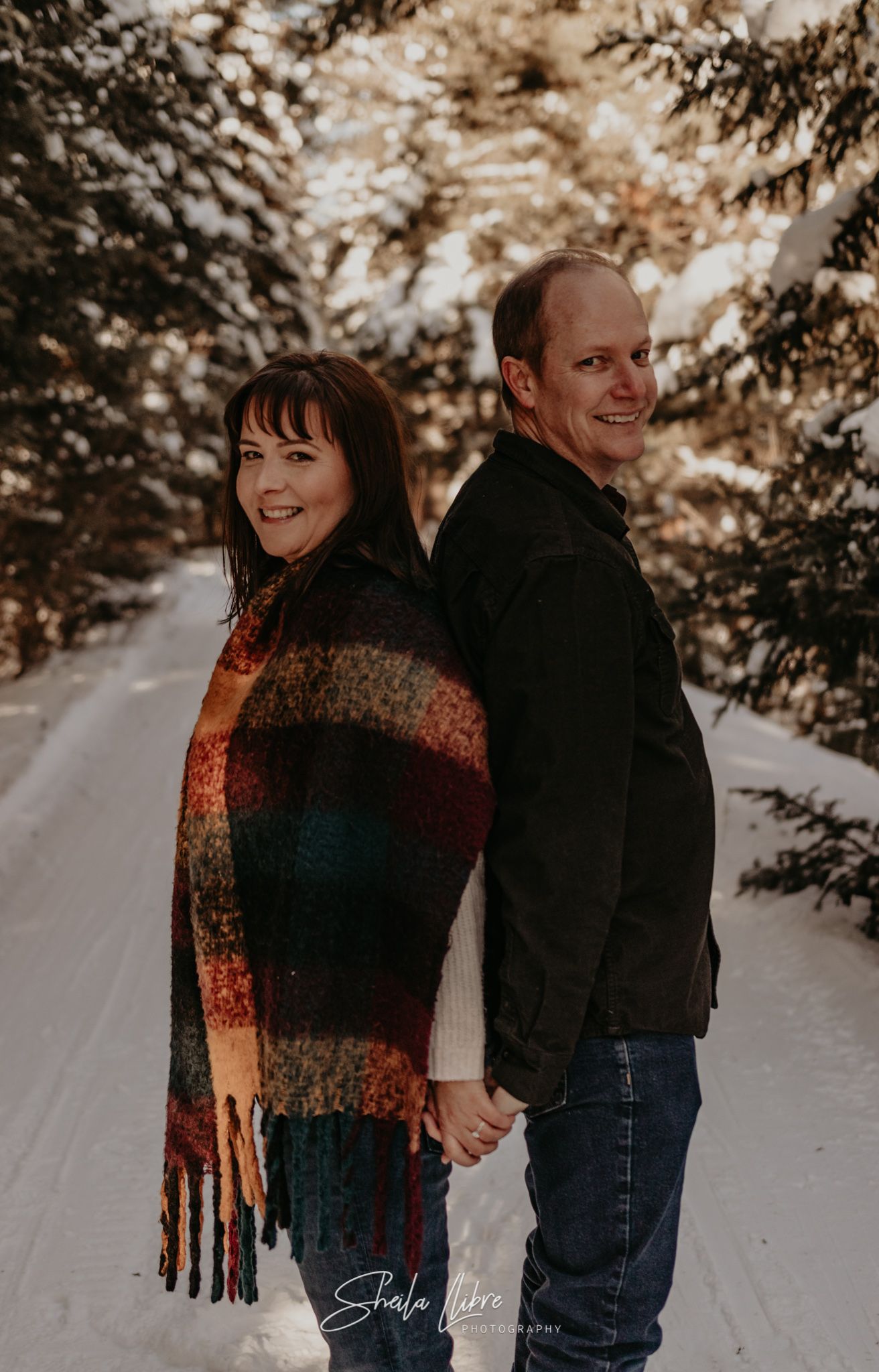 A couple holds hands and smiles while posing back-to-back on a snow-covered path.
