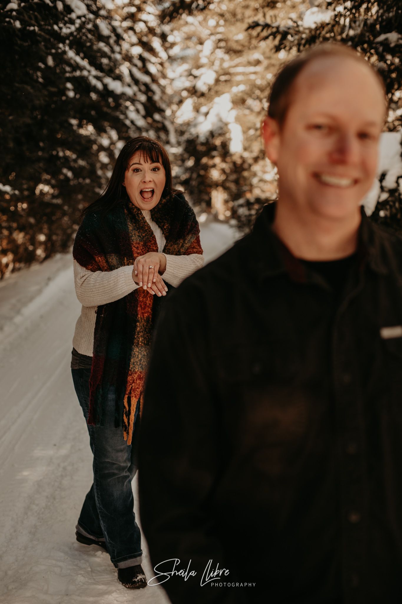 Playful winter engagement photo with one person peeking from behind the other on a snowy path.