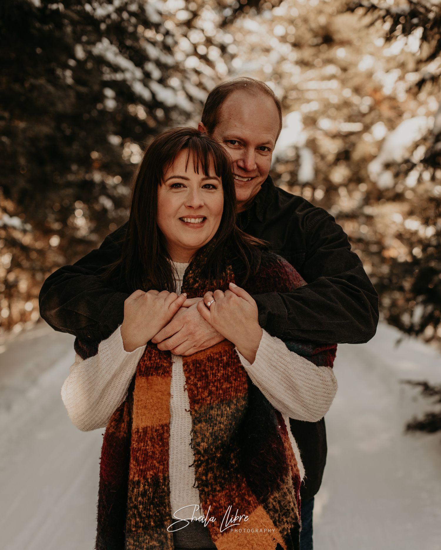 Happy winter engagement photo of couple embracing in snowy forest setting.